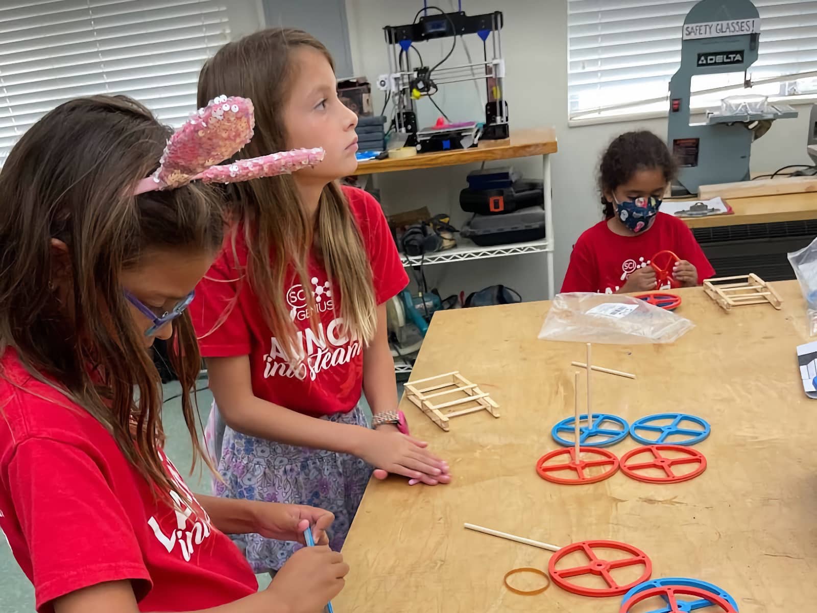 young girls at a wooden table working on a wheel and popsicle stick project