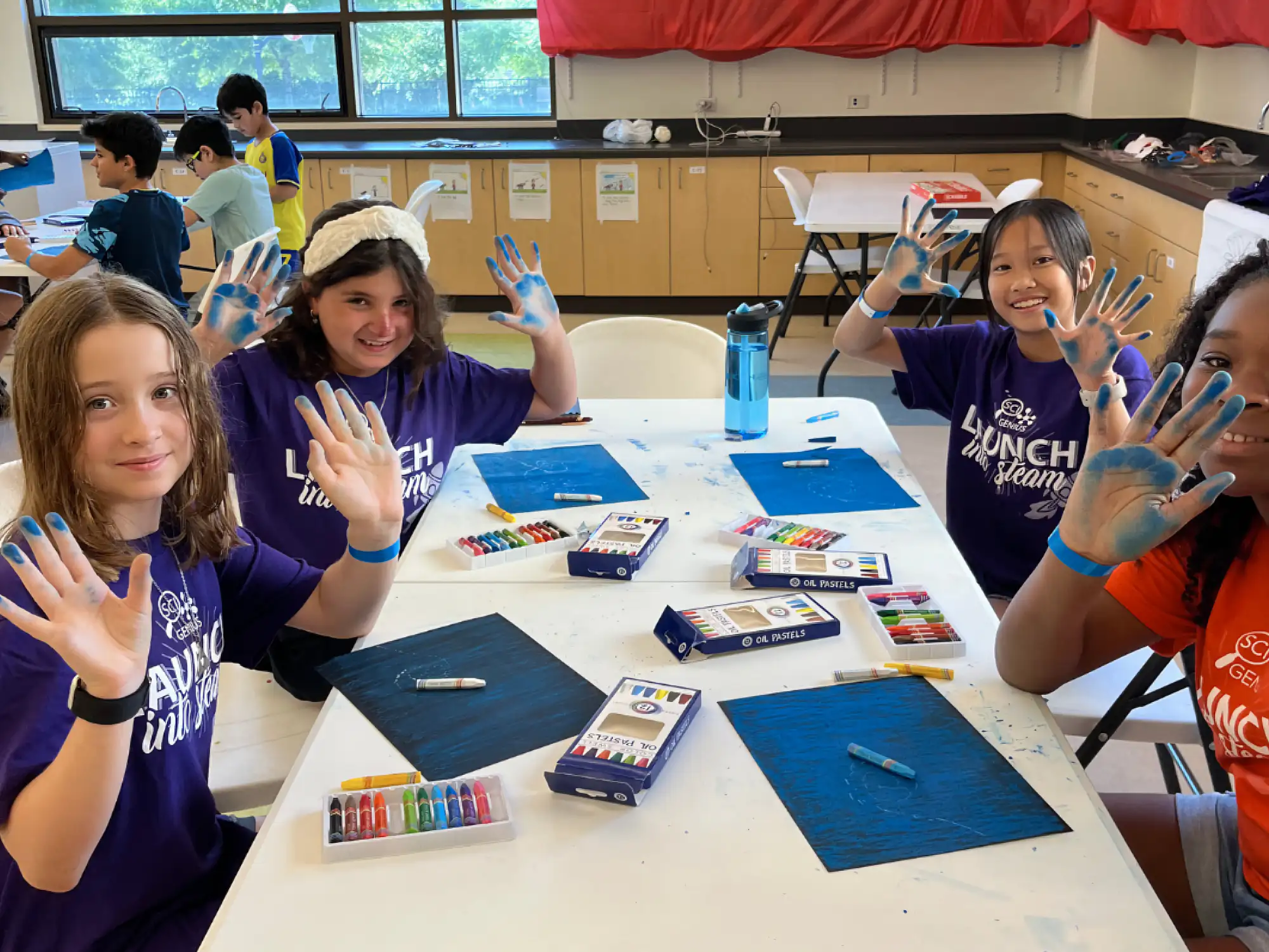 Four campers holding their blue hands up in the air while working on art projects at a table in a classroom.