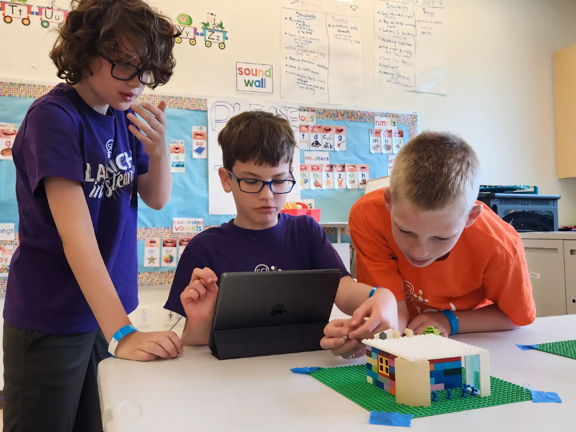Three campers using a tablet and working on a LEGO brick model in a classroom.
