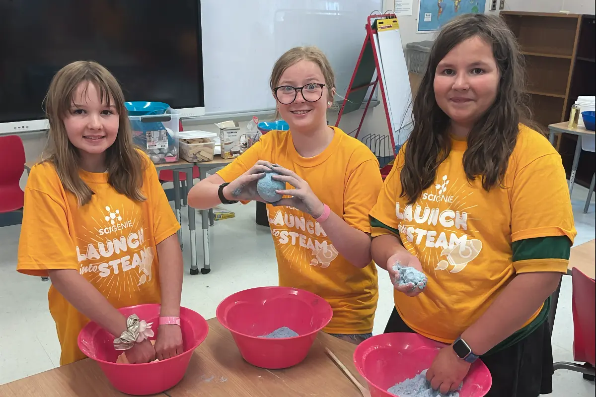 Three smiling girls in yellow SciGenie shirts mix blue material in pink bowls. The girl in the middle holds up a small blue ball.